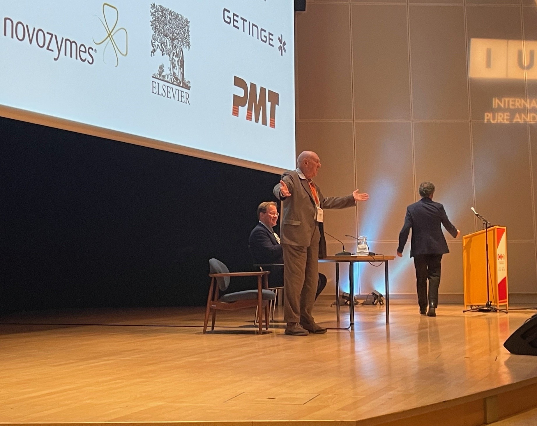 Speaker addressing audience on stage during session at European Congress on Biotechnology with sponsor logos displayed on screen